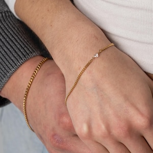 May include: Gold chain bracelets. One bracelet features a small, clear gemstone. The bracelets are worn on wrists. The image shows a person wearing a white top and blue jeans. Another person is wearing a grey jumper.