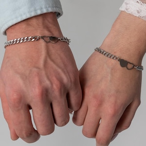 May include: Matching silver bracelets with a heart-shaped design. The bracelets are connected by a heart-shaped clasp. The bracelets are worn on wrists. The background is a solid light grey.