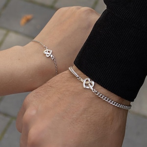 Puede incluir: Dos pulseras plateadas, cada una con un diseño de corazón. Una pulsera tiene una cadena fina, la otra una cadena de eslabones. La imagen muestra las pulseras en dos muñecas, sugiriendo un conjunto a juego.