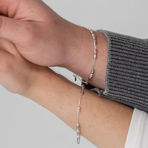 Puede incluir: Dos pulseras plateadas con pequeñas cuentas alargadas se llevan en las muñecas. Las pulseras están sobre un fondo claro. Una muñeca está cubierta por un suéter gris, y la otra lleva una camisa blanca de manga larga.