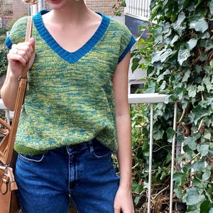 Puede incluir: Un suéter verde y azul sin mangas con cuello en V y ribete azul. El suéter está hecho de una tela suave y texturizada.