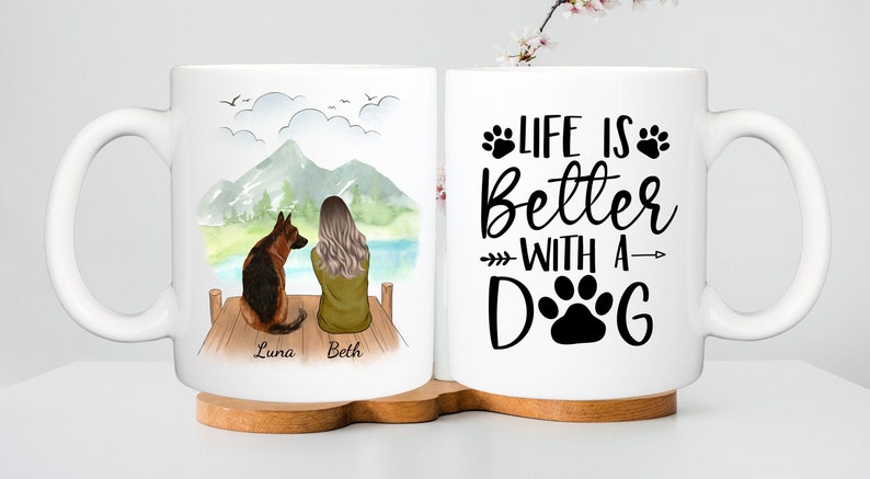 May include: White ceramic mug featuring a German Shepherd dog and a person sitting on a dock with a mountain and lake backdrop. The other side of the mug has the text "LIFE IS Better WITH A DOG" with paw prints.