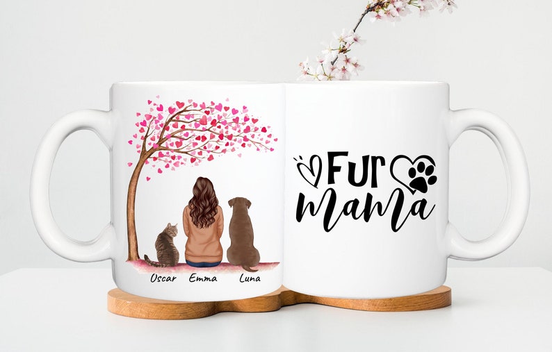 May include: White ceramic mug with a design featuring a person, a cat, and a dog under a tree with pink heart-shaped leaves. The other side says "Fur Mama" with a paw print.