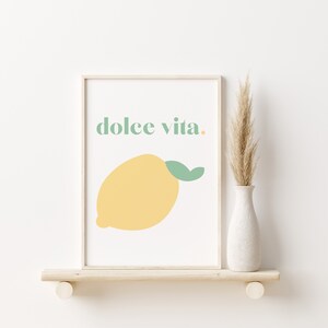 May include: A framed print with a yellow lemon and the text "dolce vita." The print is on a white background and has a light wood frame. The print is sitting on a light wood shelf.