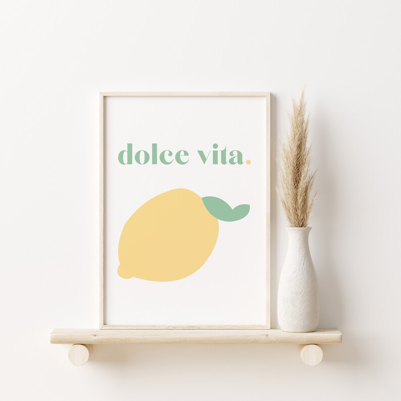 May include: A framed print with a yellow lemon and the text "dolce vita." The print is on a white background and has a light wood frame. The print is sitting on a light wood shelf.