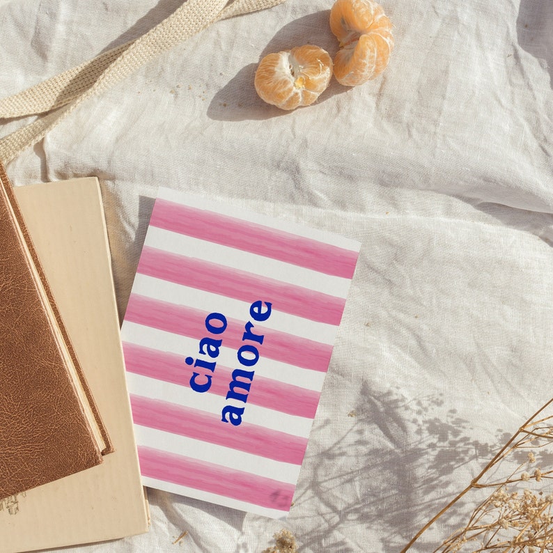 May include: A pink and white striped card with the text "ciao amore" in blue lettering. The card is lying on a white fabric surface with two peeled mandarin oranges.