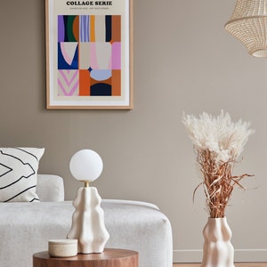 Op de afbeelding: Een ingelijste kunstdruk met de tekst "COLLAGE SERIE" en kleurrijke abstracte vormen. Een witte lamp met een ronde bol staat op een houten tafel. Een vaas met gedroogde planten en een decoratief kussen zijn ook zichtbaar.