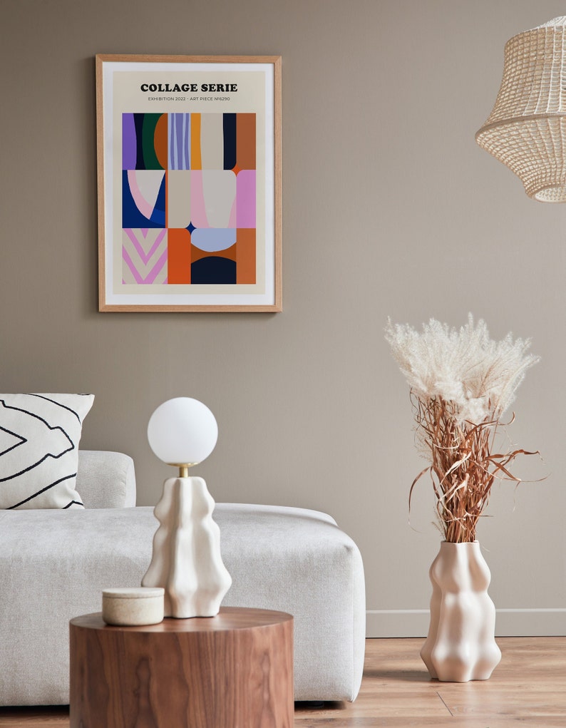 Op de afbeelding: Een ingelijste kunstdruk met de tekst "COLLAGE SERIE" en kleurrijke abstracte vormen. Een witte lamp met een ronde bol staat op een houten tafel. Een vaas met gedroogde planten en een decoratief kussen zijn ook zichtbaar.