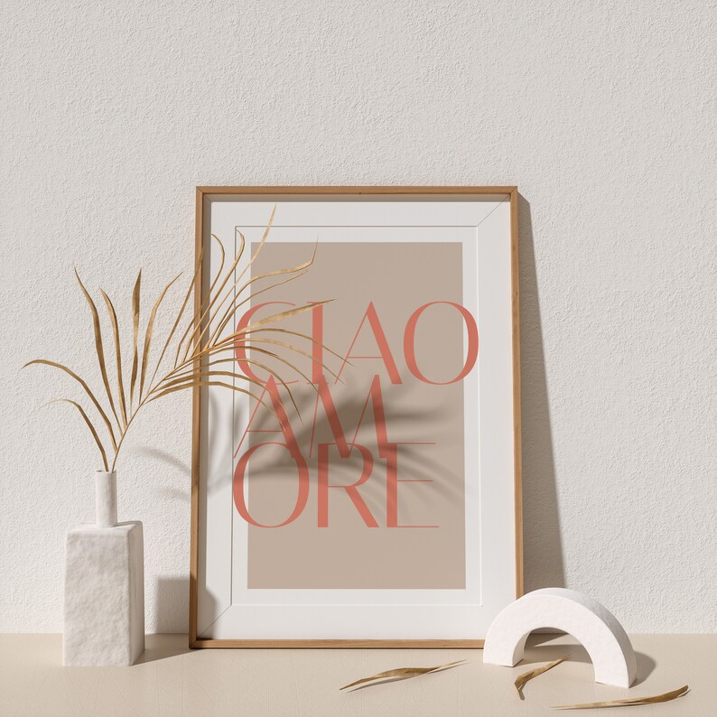 May include: A framed print with the text "CIAO AMORE" in coral pink on a beige background. The print is displayed on a white surface with a dried palm frond and a white ceramic vase.