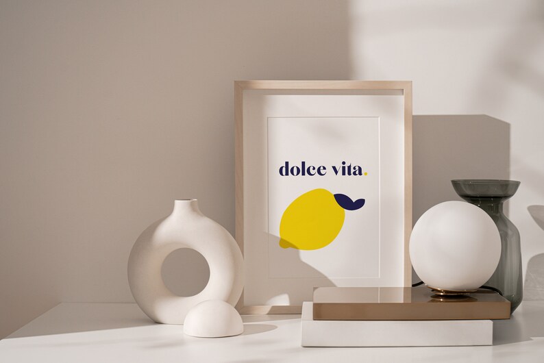 Puede incluir: Una impresi&oacute;n art&iacute;stica enmarcada con las palabras "dolce vita" y una ilustraci&oacute;n de lim&oacute;n amarillo. La impresi&oacute;n se muestra sobre una superficie blanca con jarrones decorativos y una l&aacute;mpara esf&eacute;rica. La paleta de colores incluye blanco, amarillo, azul y beige.