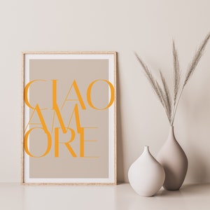 May include: A framed print with the text "CIAO AMORE" in orange letters on a beige background. The print is displayed on a white surface with two vases, one white and one beige, containing dried pampas grass.