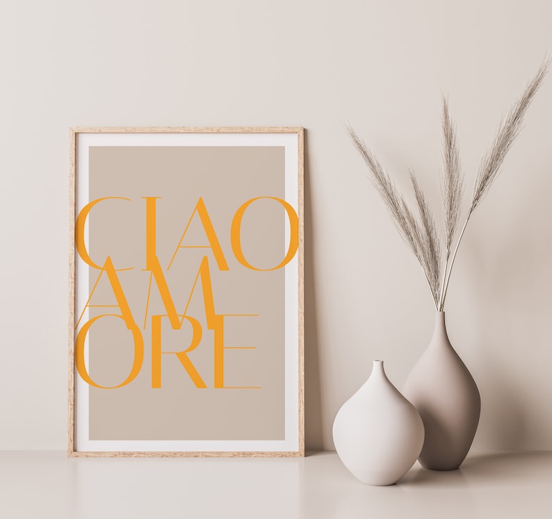 May include: A framed print with the text "CIAO AMORE" in orange letters on a beige background. The print is displayed on a white surface with two vases, one white and one beige, containing dried pampas grass.
