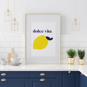 Puede incluir: Una impresi&oacute;n art&iacute;stica enmarcada con una ilustraci&oacute;n de lim&oacute;n amarillo y las palabras "dolce vita" en azul marino. La impresi&oacute;n se muestra en una cocina con azulejos blancos, l&aacute;mparas colgantes doradas y gabinetes azul marino. El lim&oacute;n tiene una hoja azul marino.