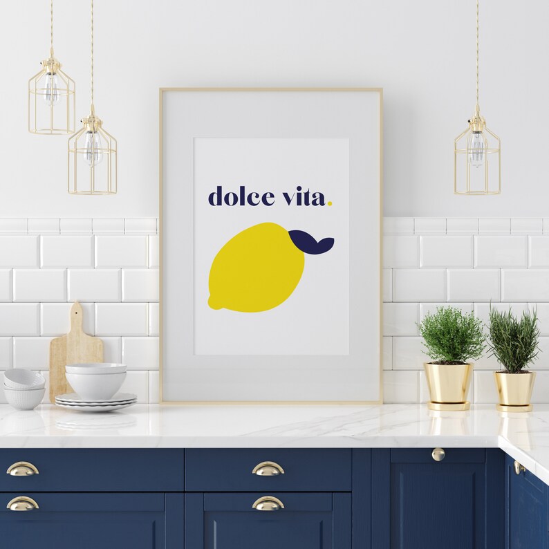 Puede incluir: Una impresi&oacute;n art&iacute;stica enmarcada con una ilustraci&oacute;n de lim&oacute;n amarillo y las palabras "dolce vita" en azul marino. La impresi&oacute;n se muestra en una cocina con azulejos blancos, l&aacute;mparas colgantes doradas y gabinetes azul marino. El lim&oacute;n tiene una hoja azul marino.