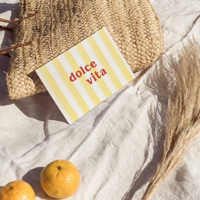 May include: A yellow and white striped card with the text "dolce vita" in red letters. The card is lying on a white fabric surface with a woven straw bag and a dried grass plume.
