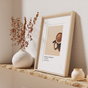 May include: A framed print of a cup of coffee with the text "Golden Morning 12.4542" and "salty brown" on a light brown shelf with a white vase with dried brown flowers and three small white ceramic vases.