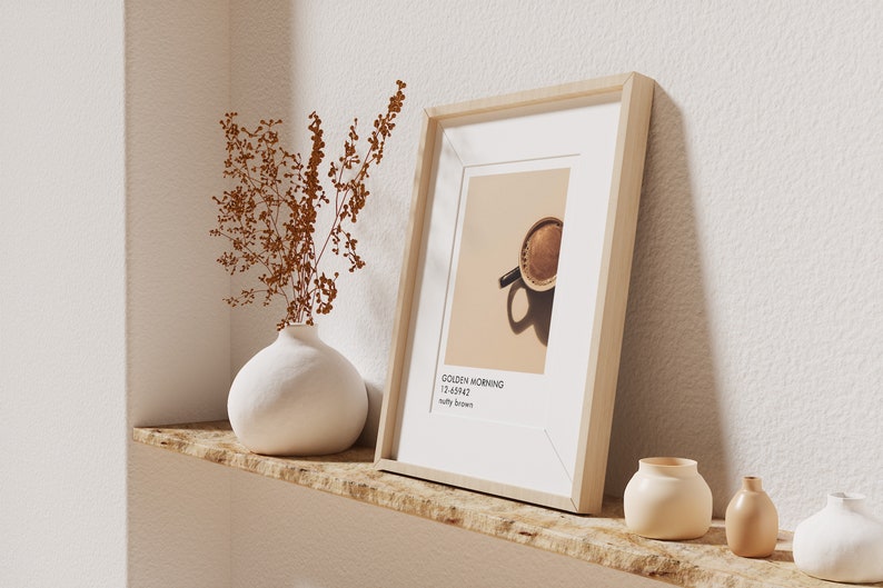 May include: A framed print of a cup of coffee with the text "Golden Morning 12.4542" and "salty brown" on a light brown shelf with a white vase with dried brown flowers and three small white ceramic vases.