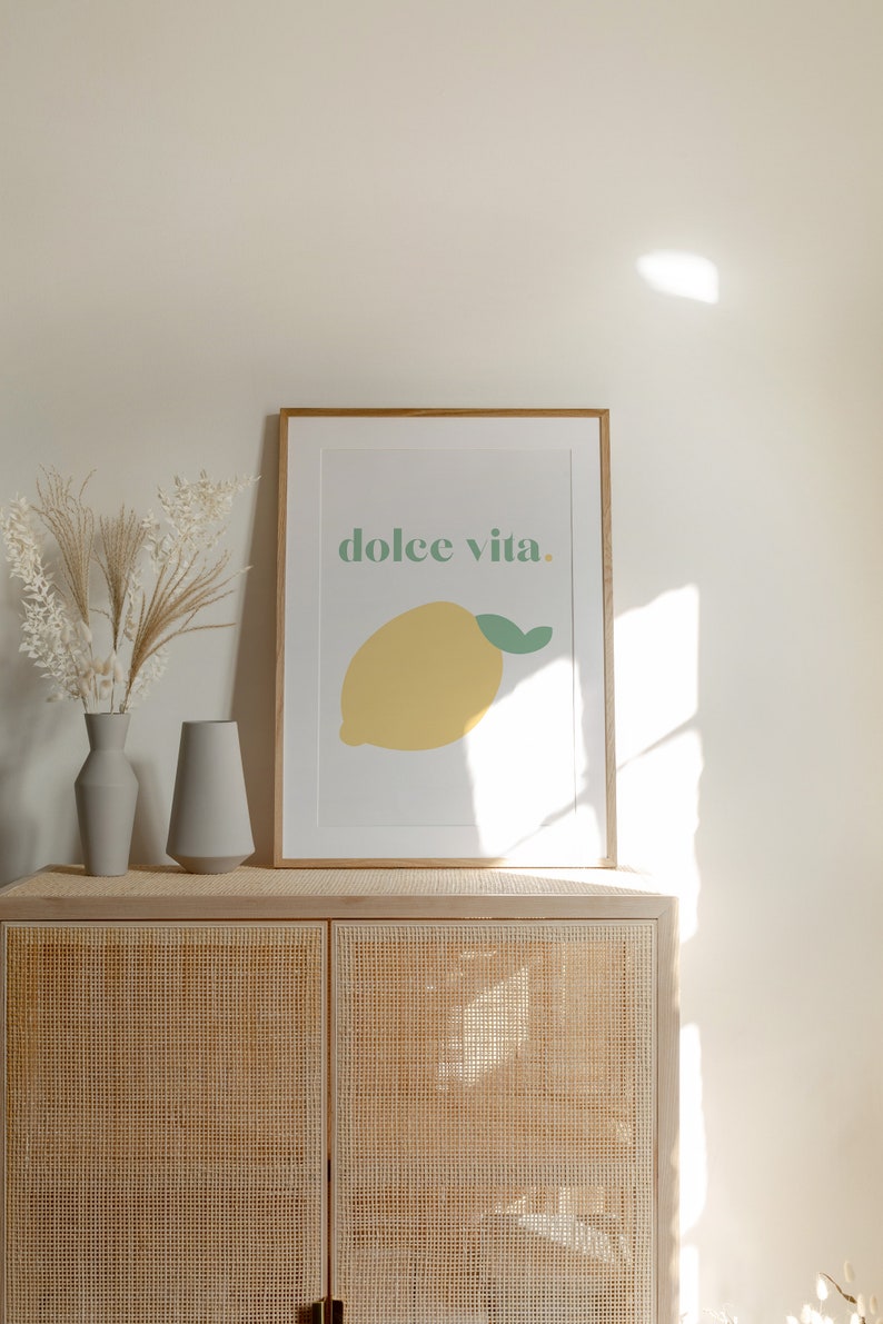 May include: A framed print with a yellow lemon and the text "dolce vita." The print is hanging on a white wall above a light brown cabinet with a woven rattan front.