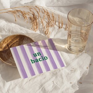 May include: A purple and white striped card with the text "un bacio" in green letters. The card is on a white fabric surface with a brown bowl and a glass of water.