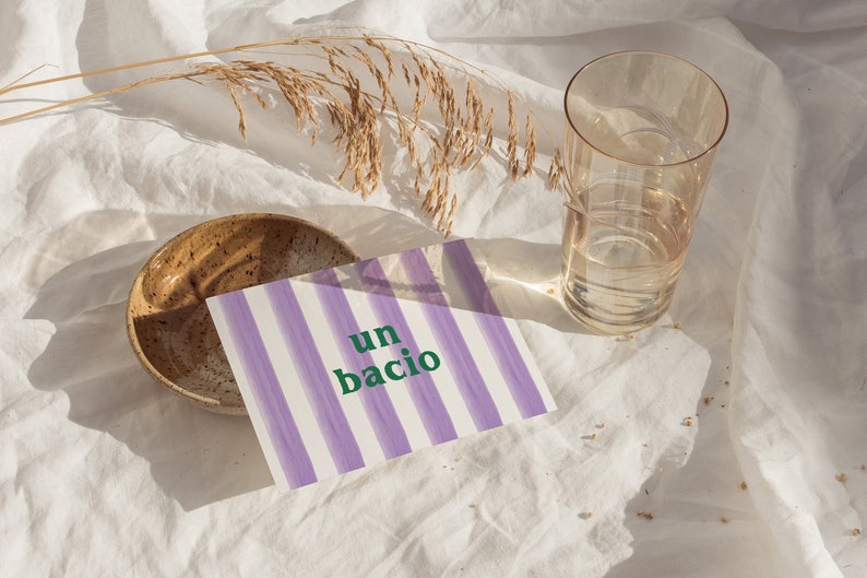 May include: A purple and white striped card with the text "un bacio" in green letters. The card is on a white fabric surface with a brown bowl and a glass of water.