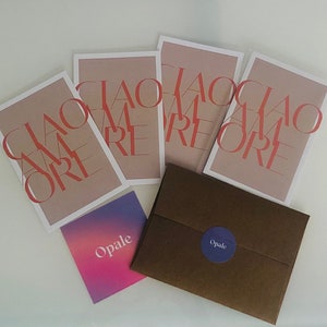 May include: Four cards with the text "Ciao Amore" in red letters on a light brown background. The cards are stacked on top of each other. A brown envelope with a purple circle with the text "Opale" is in the foreground.