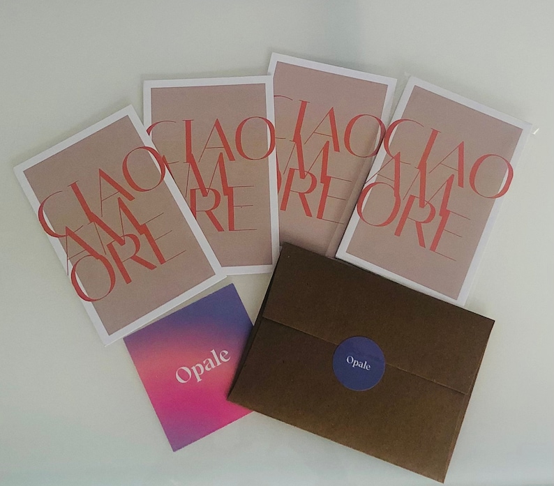 May include: Four cards with the text "Ciao Amore" in red letters on a light brown background. The cards are stacked on top of each other. A brown envelope with a purple circle with the text "Opale" is in the foreground.