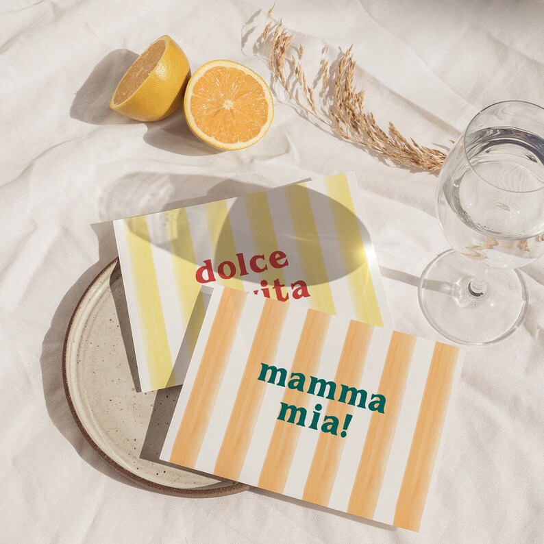 May include: Two yellow striped cards with the text "dolce vita" and "mamma mia!" on a white surface with lemons and a glass of water.