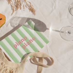 May include: A white and green striped notebook with the text "piano piano" in pink lettering. The notebook is on a white surface with a papaya, a glass of water, and a beige string bag.