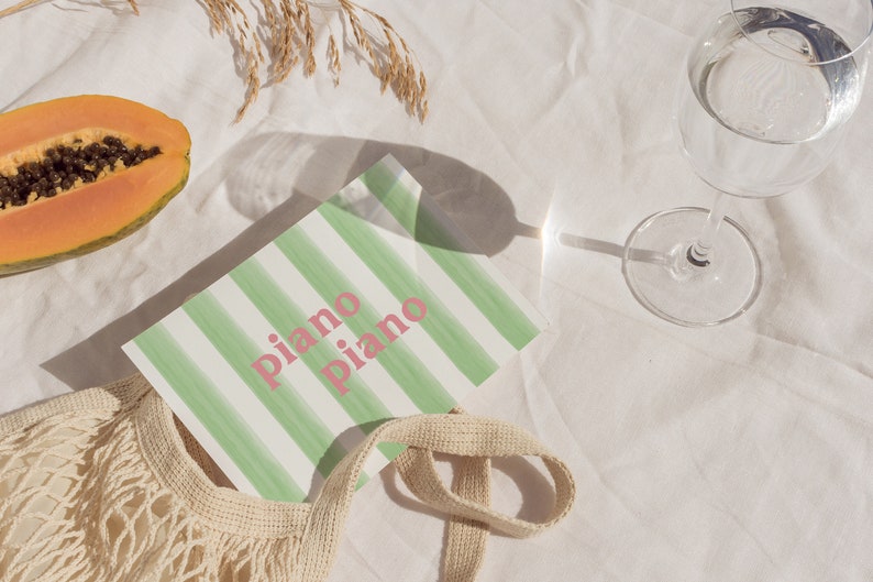 May include: A white and green striped notebook with the text "piano piano" in pink lettering. The notebook is on a white surface with a papaya, a glass of water, and a beige string bag.