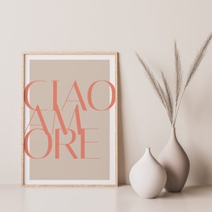 May include: A framed print with the text "CIAO AMORE" in a coral pink color on a beige background. The print is displayed on a white surface with two vases, one white and one beige, containing dried pampas grass.