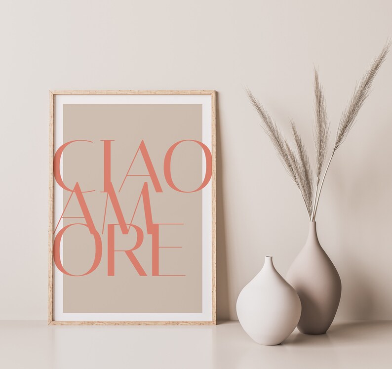 May include: A framed print with the text "CIAO AMORE" in a coral pink color on a beige background. The print is displayed on a white surface with two vases, one white and one beige, containing dried pampas grass.