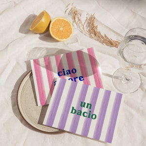 May include: Two striped cards with the words "ciao amore" and "un bacio" written in blue and green lettering. The cards are on a white surface with lemons and a glass of water.