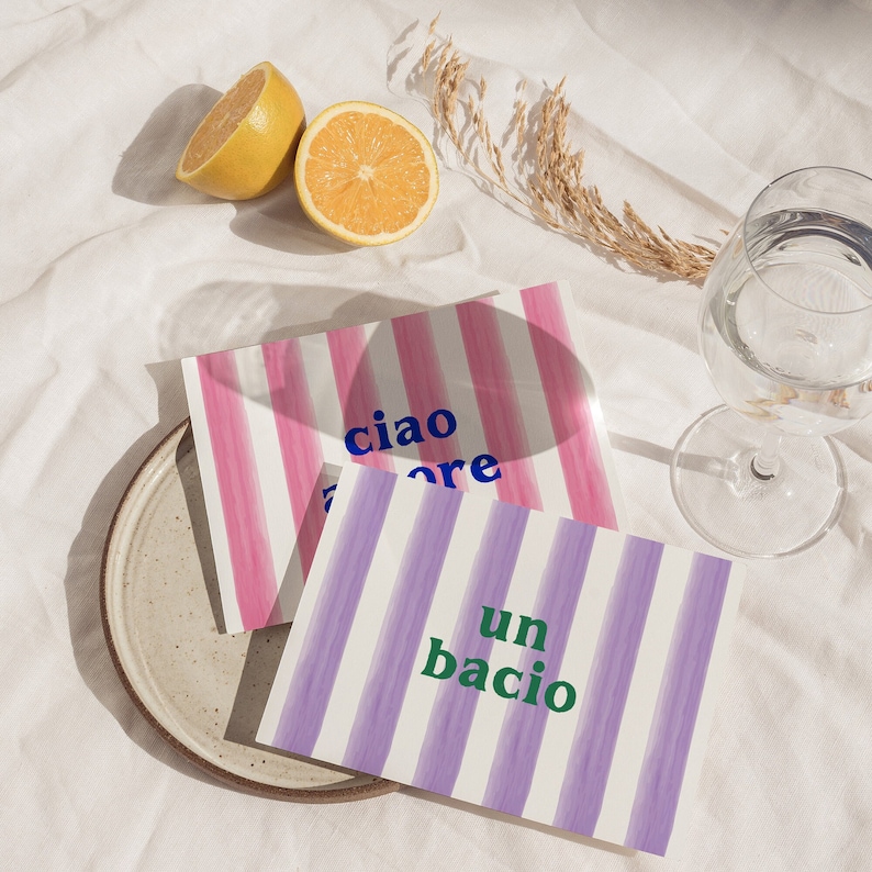 May include: Two striped cards with the words "ciao amore" and "un bacio" written in blue and green lettering. The cards are on a white surface with lemons and a glass of water.
