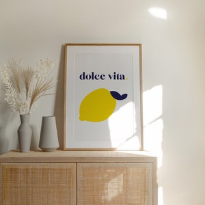 Puede incluir: Una impresi&oacute;n art&iacute;stica enmarcada con un lim&oacute;n amarillo y una hoja azul marino, con las palabras "dolce vita" en azul marino. La impresi&oacute;n est&aacute; sobre un mueble claro con dos jarrones grises y flores secas. El mueble es de material tejido.