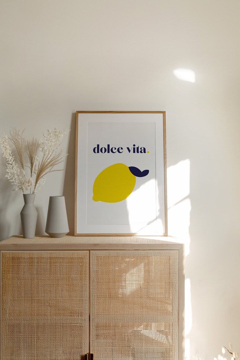 Puede incluir: Una impresi&oacute;n art&iacute;stica enmarcada con un lim&oacute;n amarillo y una hoja azul marino, con las palabras "dolce vita" en azul marino. La impresi&oacute;n est&aacute; sobre un mueble claro con dos jarrones grises y flores secas. El mueble es de material tejido.
