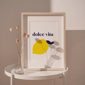 Puede incluir: Una impresi&oacute;n art&iacute;stica enmarcada con una ilustraci&oacute;n de lim&oacute;n amarillo y una hoja azul marino, con las palabras "dolce vita". La impresi&oacute;n est&aacute; en un marco de madera clara. Un jarr&oacute;n con flores secas est&aacute; sobre una mesa blanca junto a la impresi&oacute;n enmarcada.