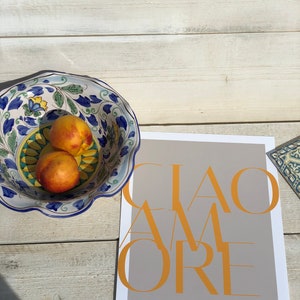May include: A grey card with the text "CIAO AMORE" in yellow letters. A blue and white ceramic bowl with peaches sits on a white wooden surface.