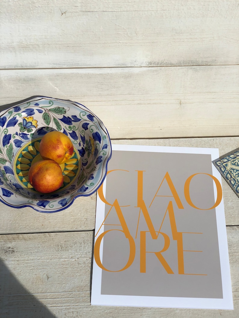 May include: A grey card with the text "CIAO AMORE" in yellow letters. A blue and white ceramic bowl with peaches sits on a white wooden surface.