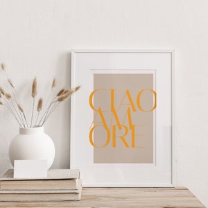 May include: A white framed print with the text "CIAO AMORE" in orange on a light brown background. The print is sitting on a wooden table with a stack of books and a vase with dried flowers.