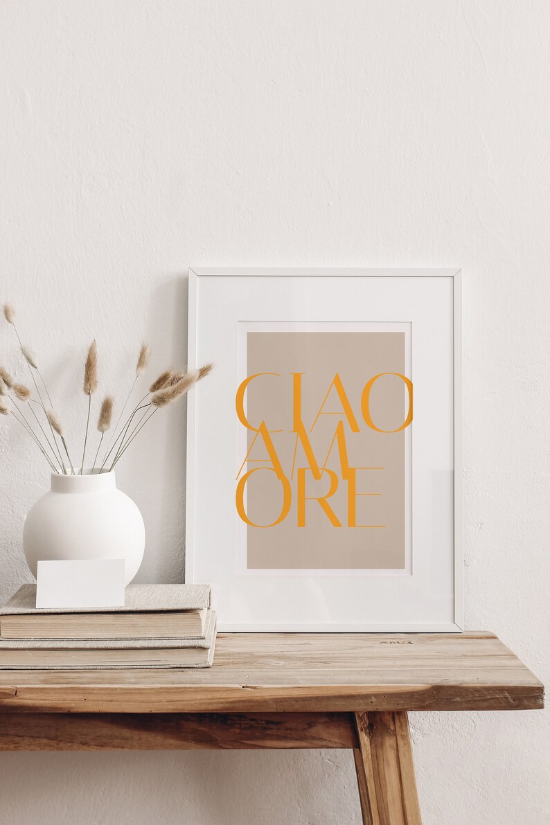 May include: A white framed print with the text "CIAO AMORE" in orange on a light brown background. The print is sitting on a wooden table with a stack of books and a vase with dried flowers.