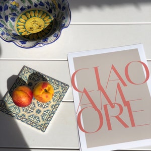 May include: A white ceramic bowl with a blue and yellow floral design, two peaches on a blue and white tiled tray, and a beige poster with the text "CIAO AMORE" in pink.