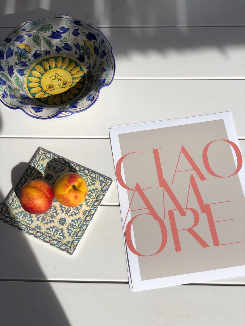 May include: A white ceramic bowl with a blue and yellow floral design, two peaches on a blue and white tiled tray, and a beige poster with the text "CIAO AMORE" in pink.