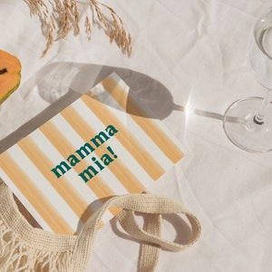 May include: A white and orange striped card with the text "mamma mia!" in green letters. The card is lying on a white tablecloth with a papaya, a mesh bag, and a glass of water.
