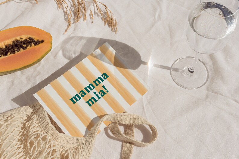 May include: A white and orange striped card with the text "mamma mia!" in green letters. The card is lying on a white tablecloth with a papaya, a mesh bag, and a glass of water.