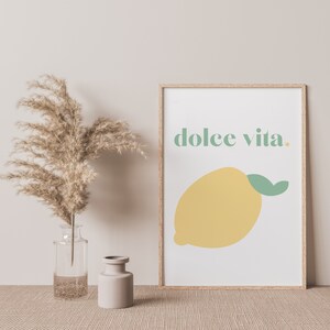 May include: A framed print with a white background and a yellow lemon with a green leaf. The text "dolce vita." is written in green letters above the lemon.