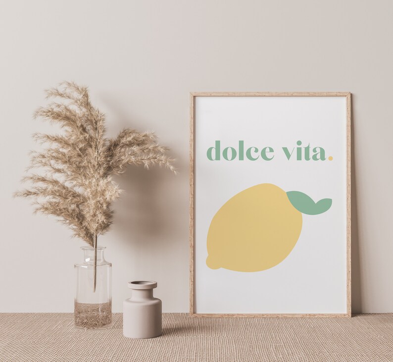 May include: A framed print with a white background and a yellow lemon with a green leaf. The text "dolce vita." is written in green letters above the lemon.