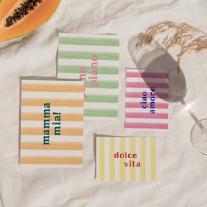May include: Four colorful striped cards with Italian phrases on a white fabric background. The phrases are "mamma mia!" in green and orange, "dolce vita" in red and yellow, "ciao amore" in blue and pink, and "no piano" in pink and green.