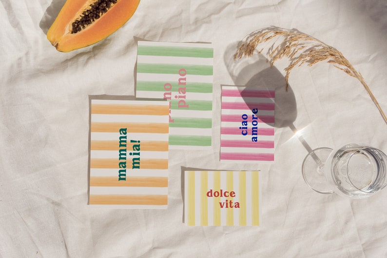 May include: Four colorful striped cards with Italian phrases on a white fabric background. The phrases are "mamma mia!" in green and orange, "dolce vita" in red and yellow, "ciao amore" in blue and pink, and "no piano" in pink and green.