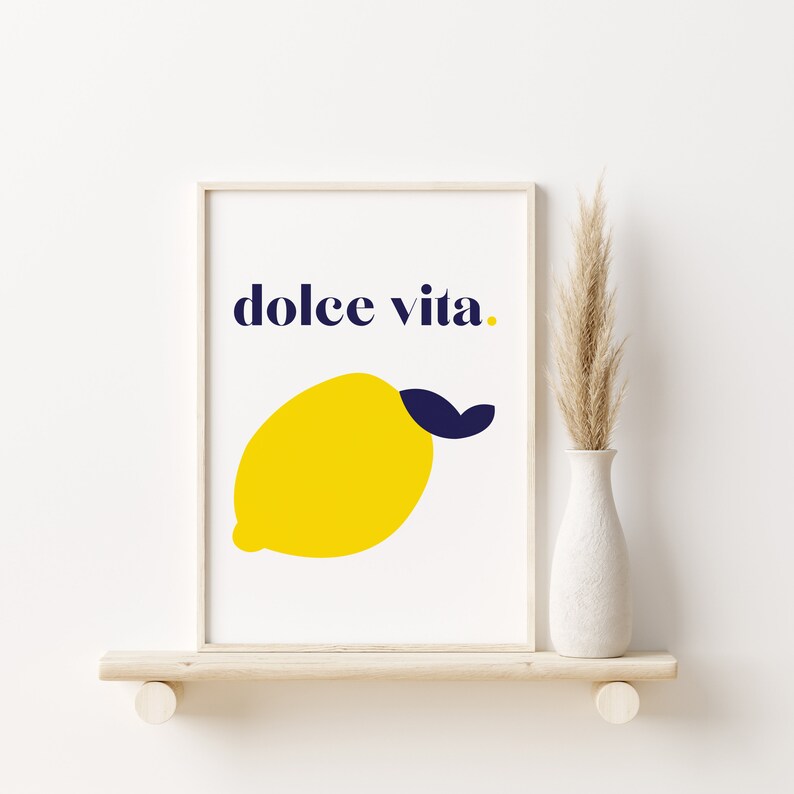 Puede incluir: Una impresi&oacute;n art&iacute;stica enmarcada con una ilustraci&oacute;n de lim&oacute;n amarillo y una hoja azul marino sobre un fondo blanco. Las palabras "dolce vita" est&aacute;n escritas encima del lim&oacute;n en azul marino. La impresi&oacute;n se exhibe en una estanter&iacute;a de madera con un jarr&oacute;n blanco y plantas secas.