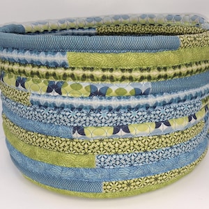 May include: A round, fabric storage basket with a blue and green patchwork design. The basket features a variety of patterned fabrics in shades of blue and green, with rope handles on each side. The basket is approximately 30 cm in diameter and 20 cm tall.